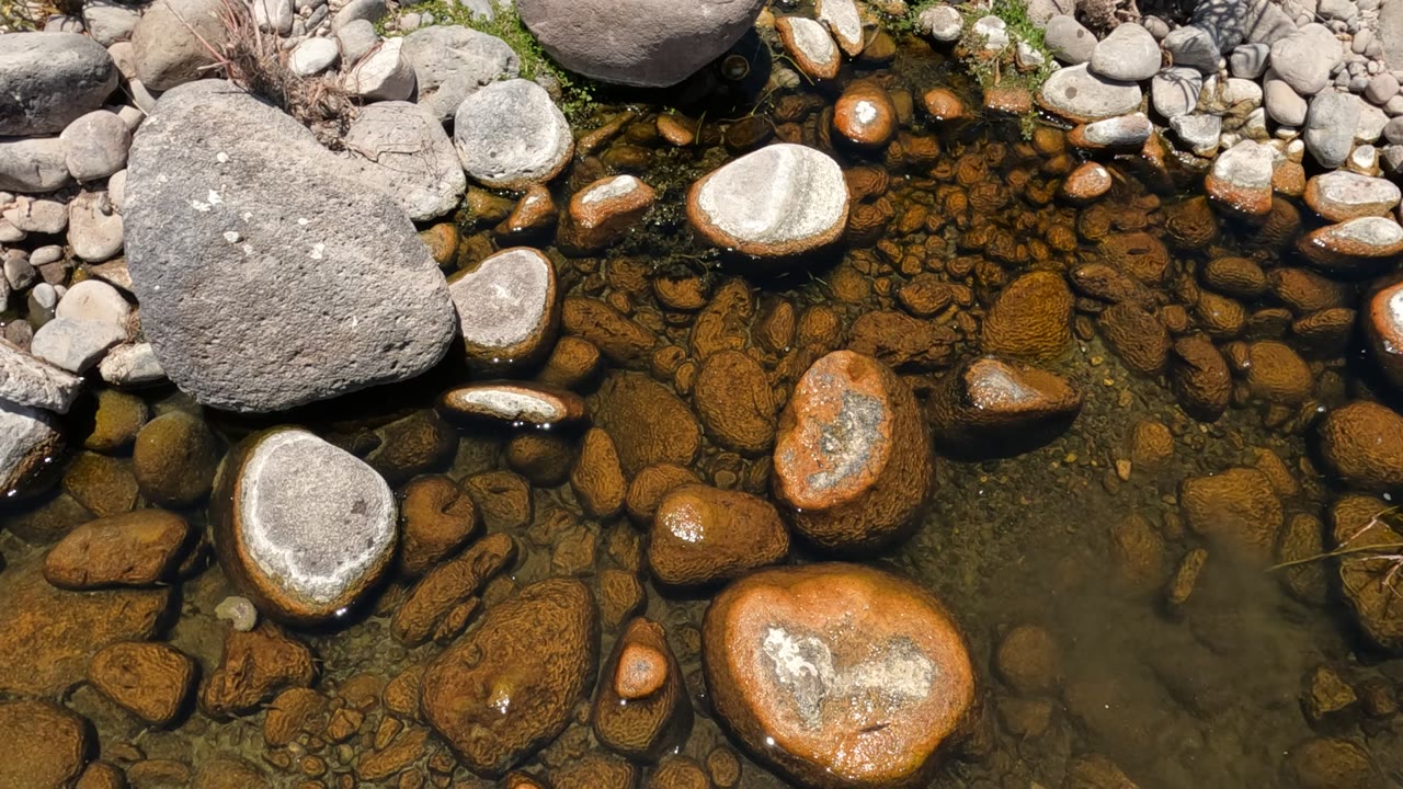 Clear shallow stream water in Baja California, Mexico flows over brown and gray rounded rocks with a large pale stone at upper left and no visible people.