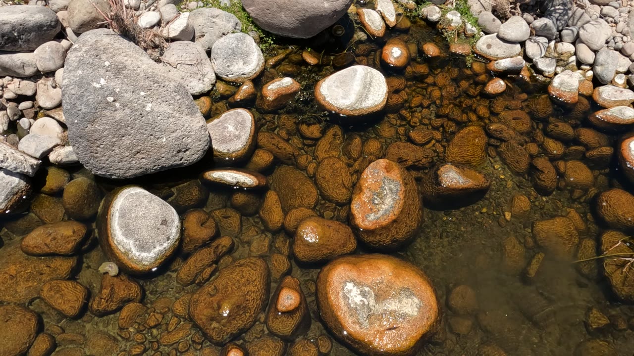 A close downward view in Baja California, Mexico shows clear stream water passing over rounded orange-brown stones and gray pebbles, with no visible people.