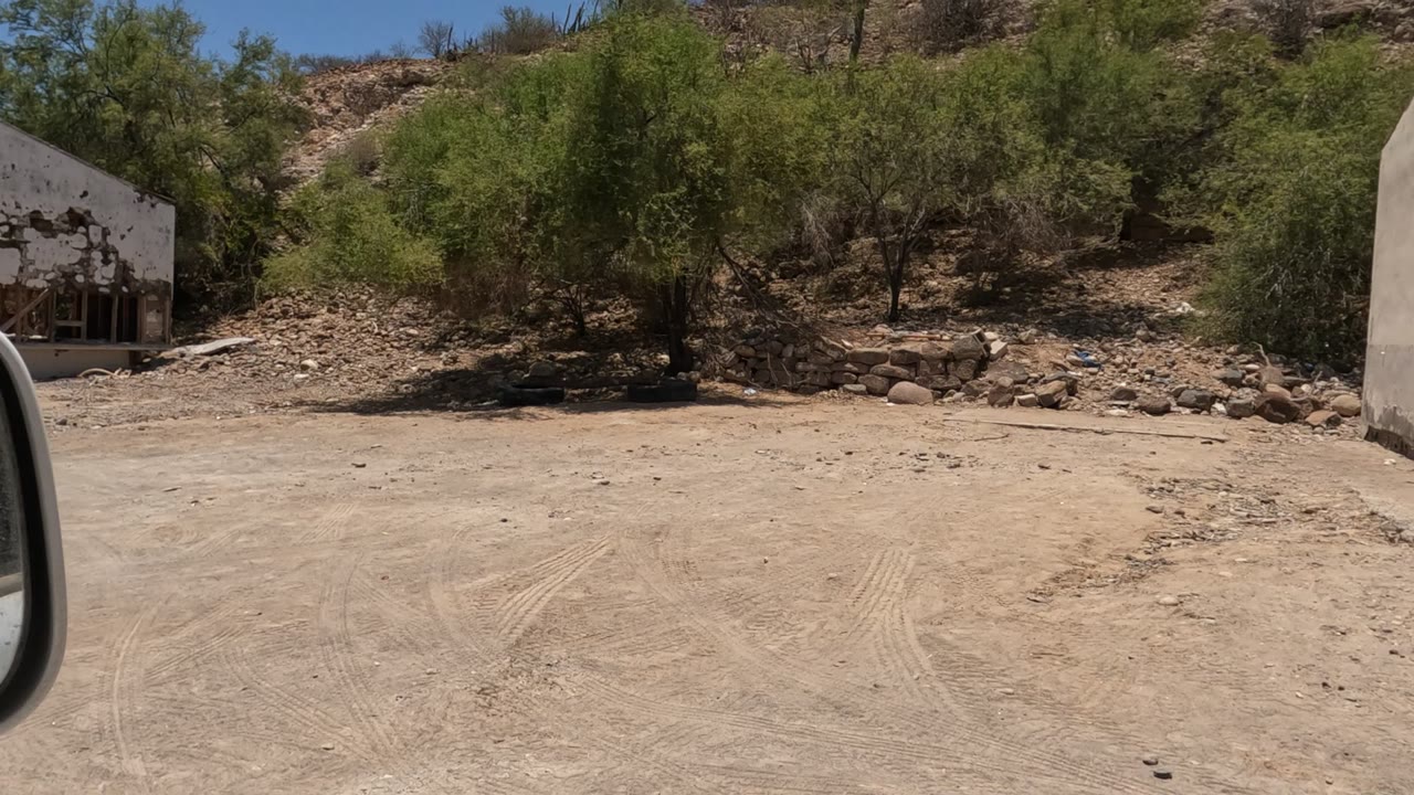 Early frame in Baja California showing a dirt roadside, rocky slope, green desert bushes, and the edge of a damaged white building, with no visible people.