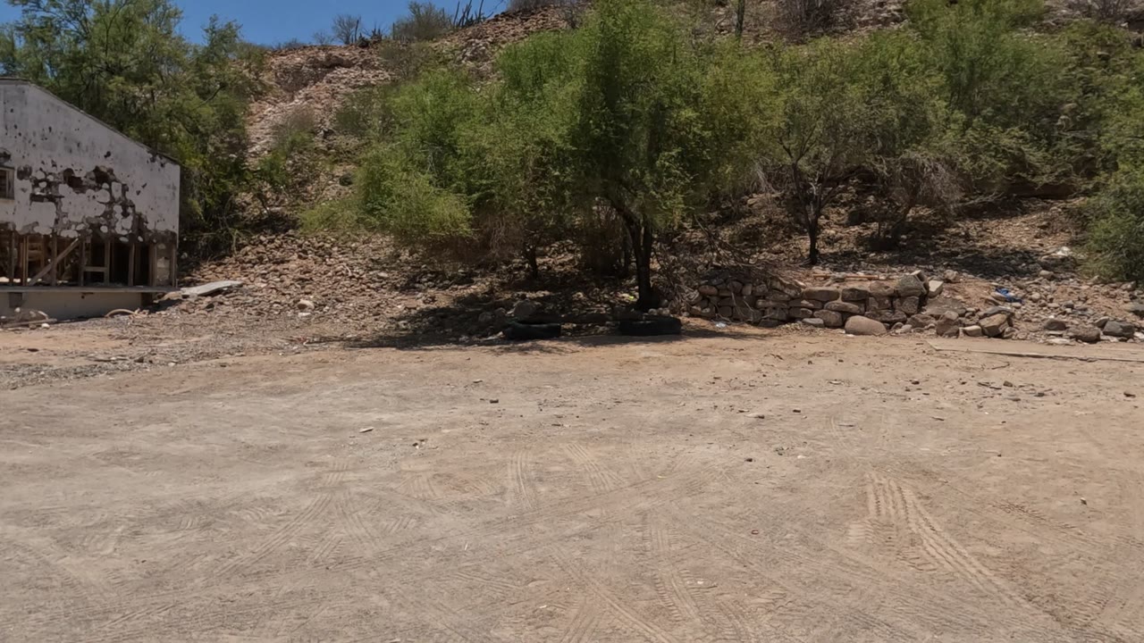 Baja California roadside view of a small deteriorated white building set against green brush and a rocky hillside, with no visible people.