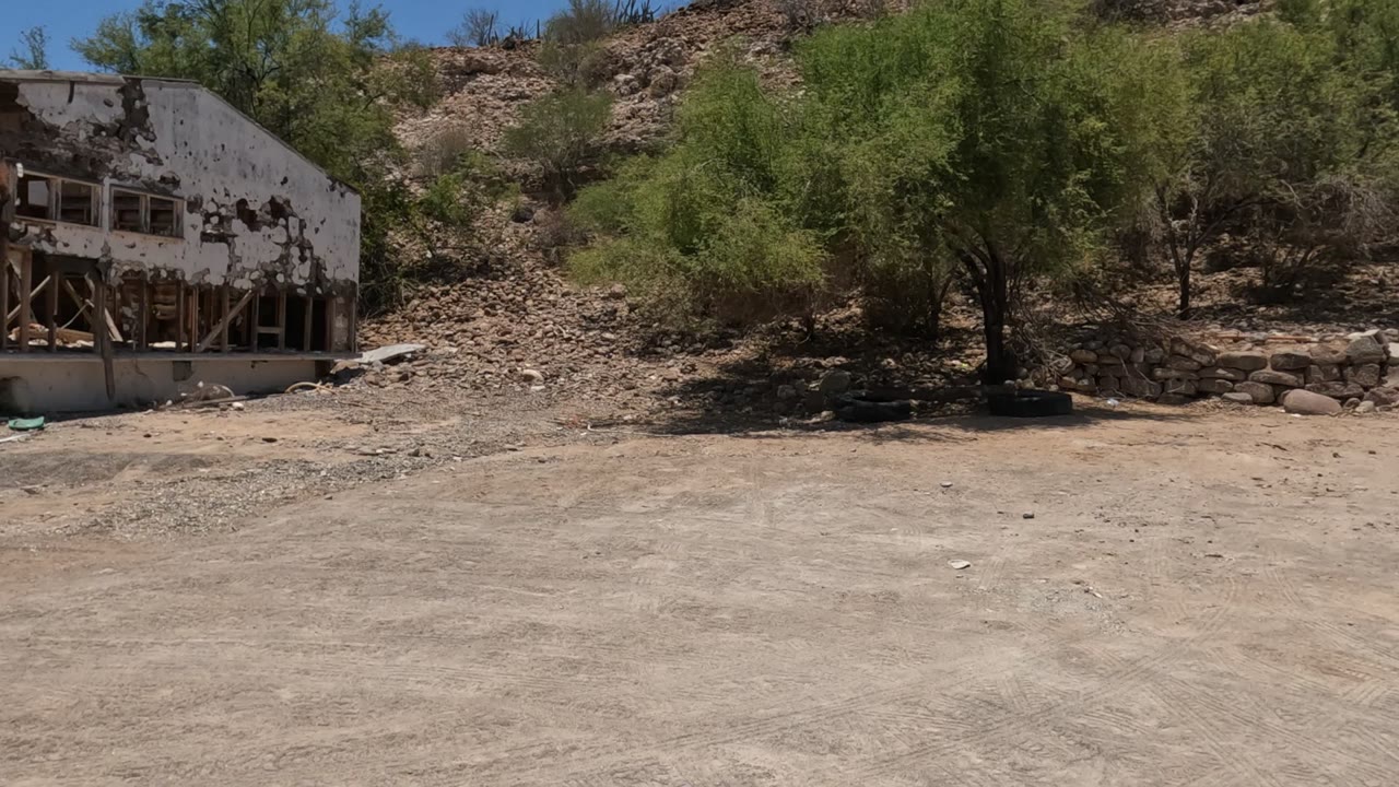 Closer Baja California frame showing the ruined building front with broken wall sections, exposed wood framing, rubble, bushes, and no visible people.