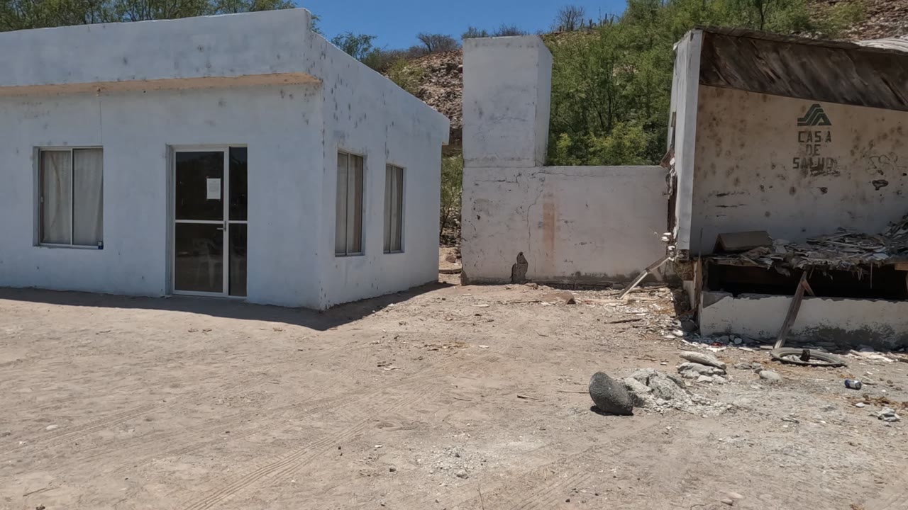 At the start of the clip, a small white building in Baja California stands on dusty open ground with windows, a glass door, dry hillside, and no visible people.