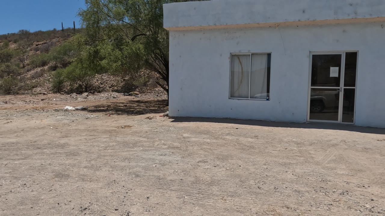 Midway through the pan, the white concrete building in Baja California is framed more frontally with a window, glass door, dirt yard, hillside, and no visible people.