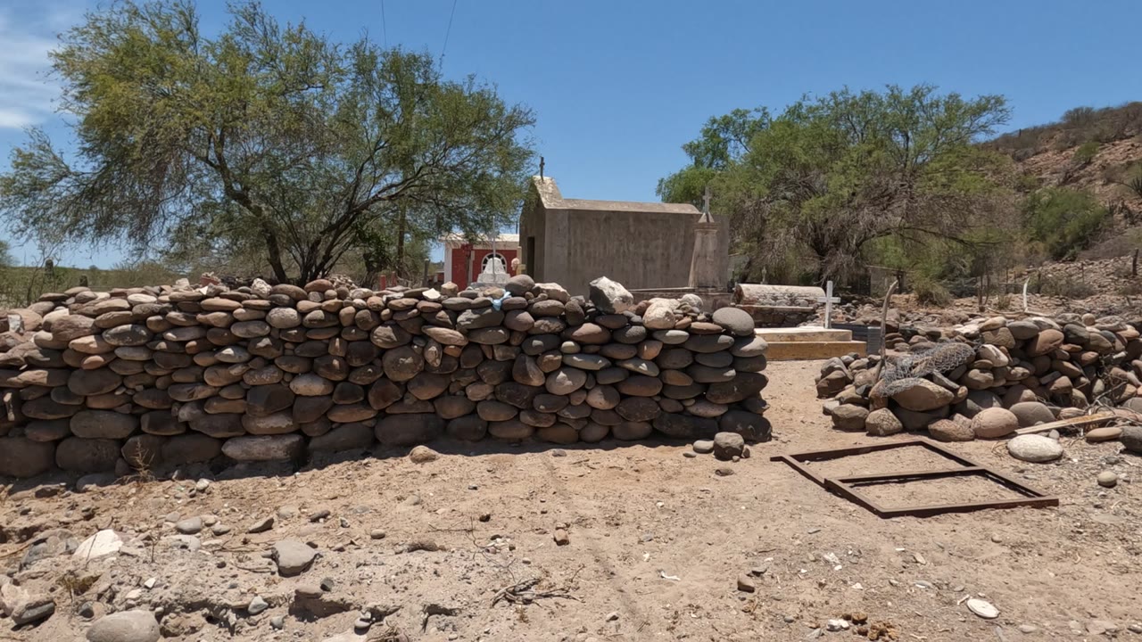 Baja California, Mexico frame showing a low stacked stone wall across dry ground, trees, a hillside, and a small structure with cross-shaped markers in the background.