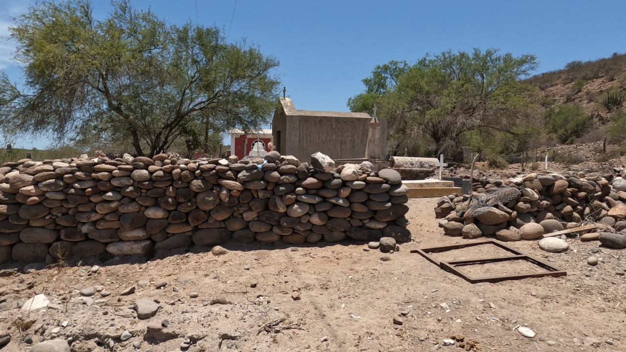Rounded stone wall with scattered rocks, trees, hillside, and a small structure behind.