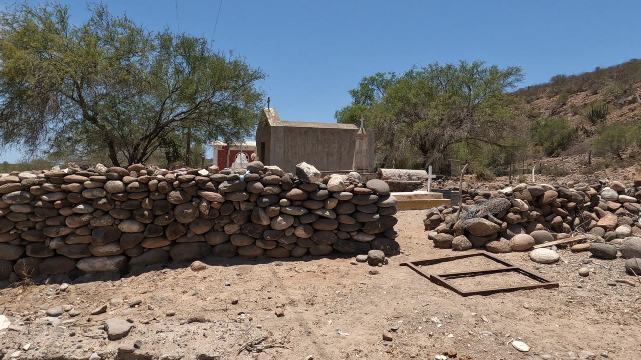 Baja California, Mexico frame showing the stone wall running across the foreground, a rectangular metal frame on dusty ground, trees, hillside, and a small structure behind.
