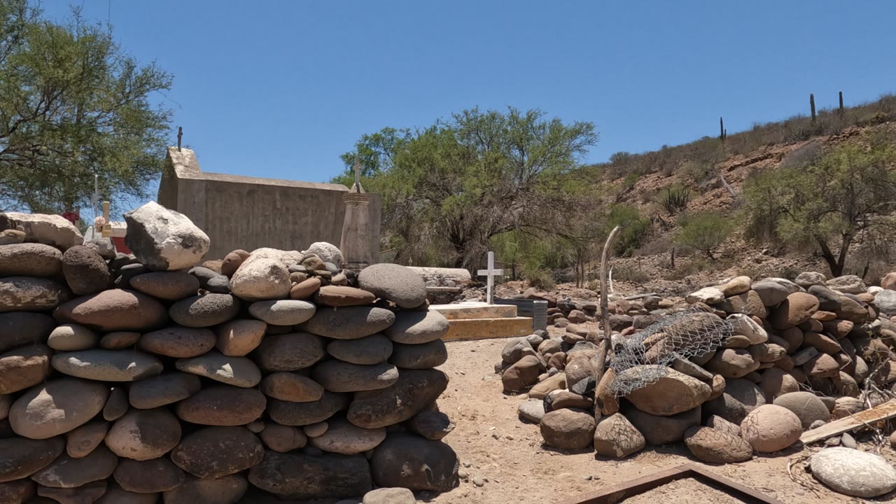 Opening frame in Baja California, Mexico showing a rounded stone wall in the foreground, a small chapel structure behind it, dry ground, trees, rocky hillside, blue sky, and no visible people.