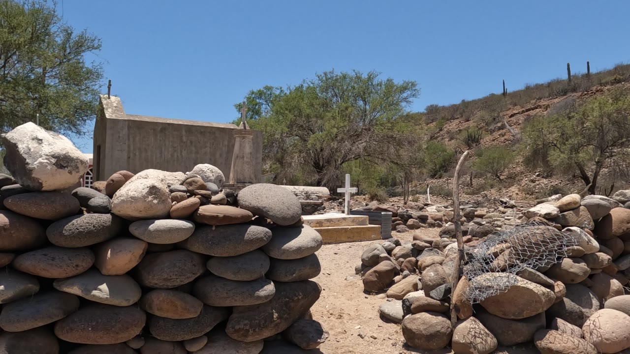 Stone wall and white cross with a chapel wall behind them in dry terrain.