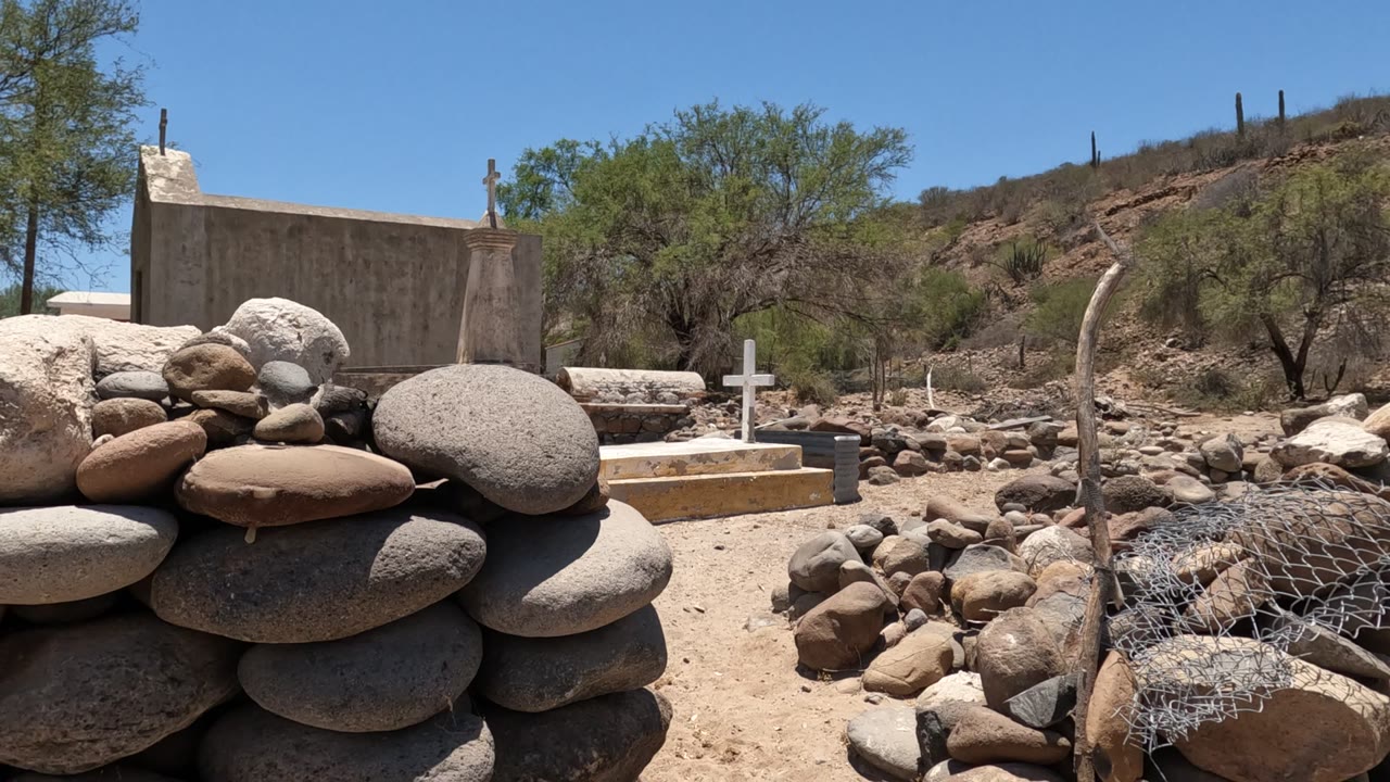 Baja California, Mexico frame with large rounded rocks filling the foreground, white crosses and low grave markers behind them, a chapel wall, sparse green trees, rocky slope, blue sky, and no visible people.