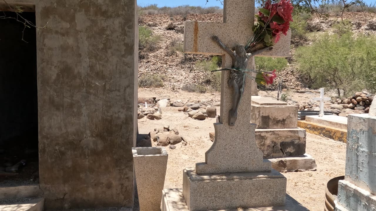 A stone crucifix with red flowers stands near above-ground cemetery tombs in Baja California, Mexico, with rocky ground, sparse shrubs, and no visible people.