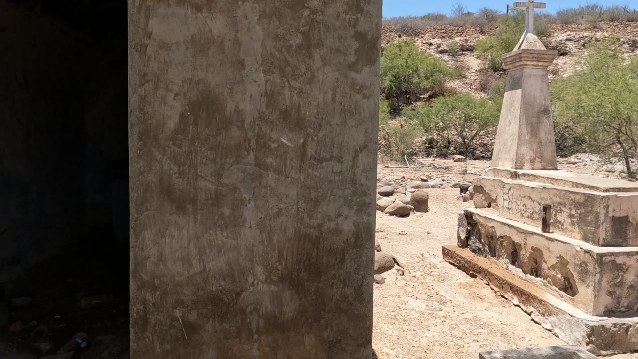A sunlit tomb with a white cross appears beyond a rough concrete wall in Baja California, Mexico, with dry ground and no visible people.