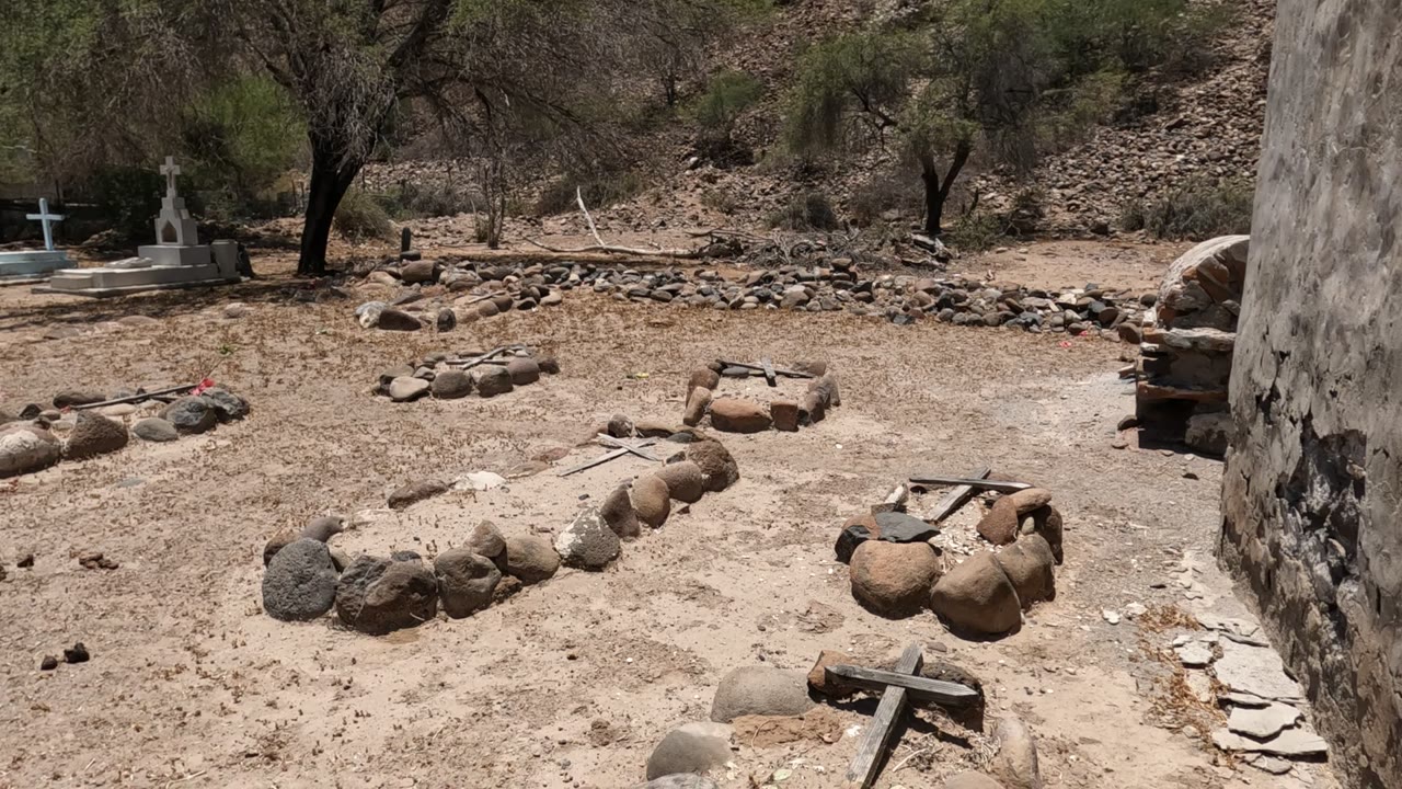 Dry cemetery with low stone graves, crosses, sparse trees, and rocky hillside.