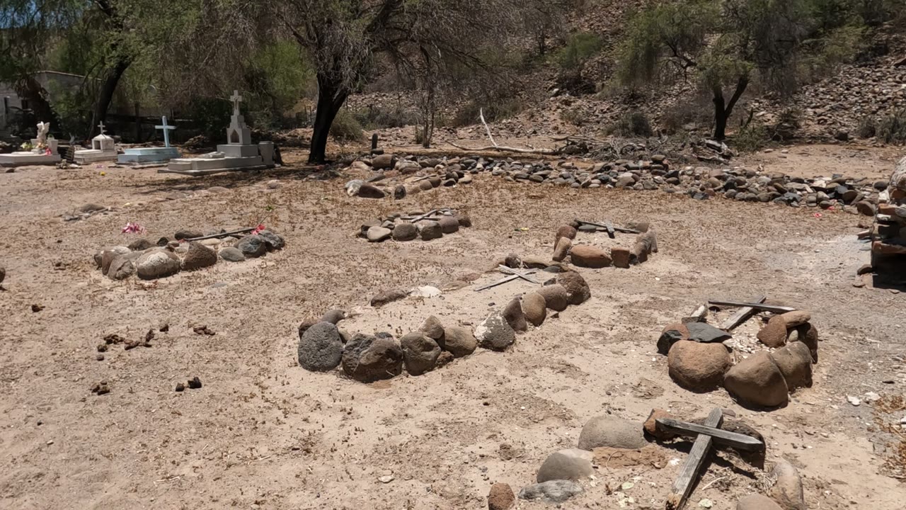 Baja California cemetery frame with a rough stone wall line, multiple stone-ringed graves, white crosses near the back, dry hillside, and no visible people.