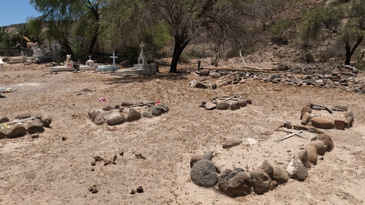 A Baja California cemetery frame shows low stone grave outlines in sandy ground, a red-brick chapel wall at right, crosses and tomb markers in the background, and no visible people.