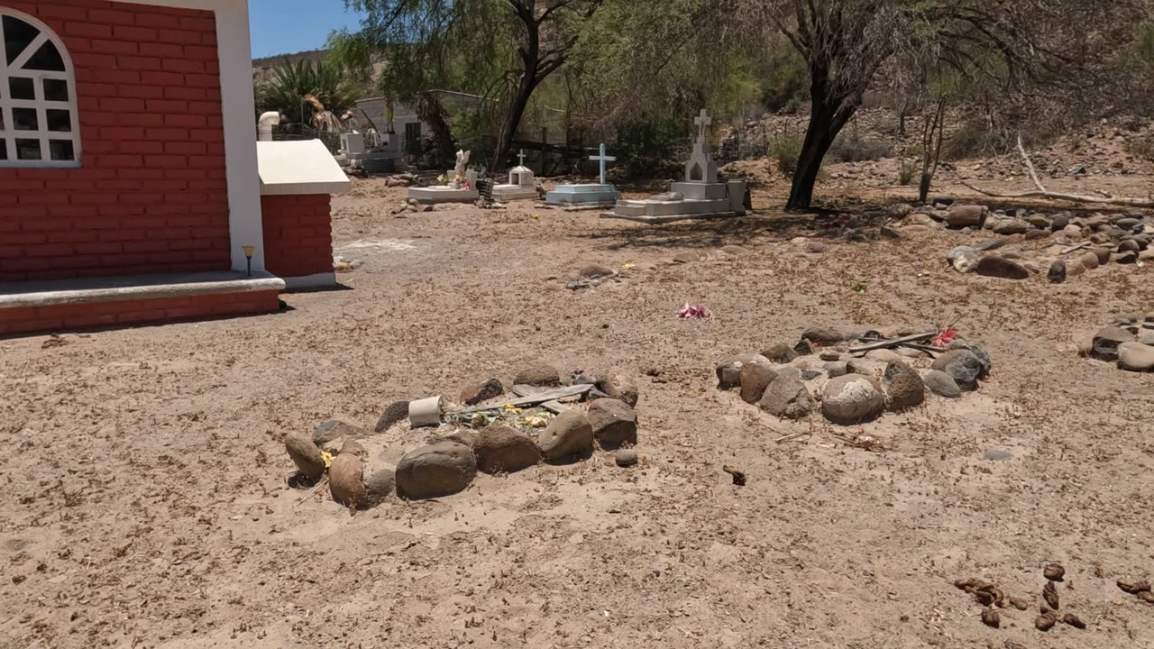 A later Baja California cemetery frame centers the sandy burial ground with stone grave borders in front, white crosses and tomb markers behind, trees overhead, and no visible people.