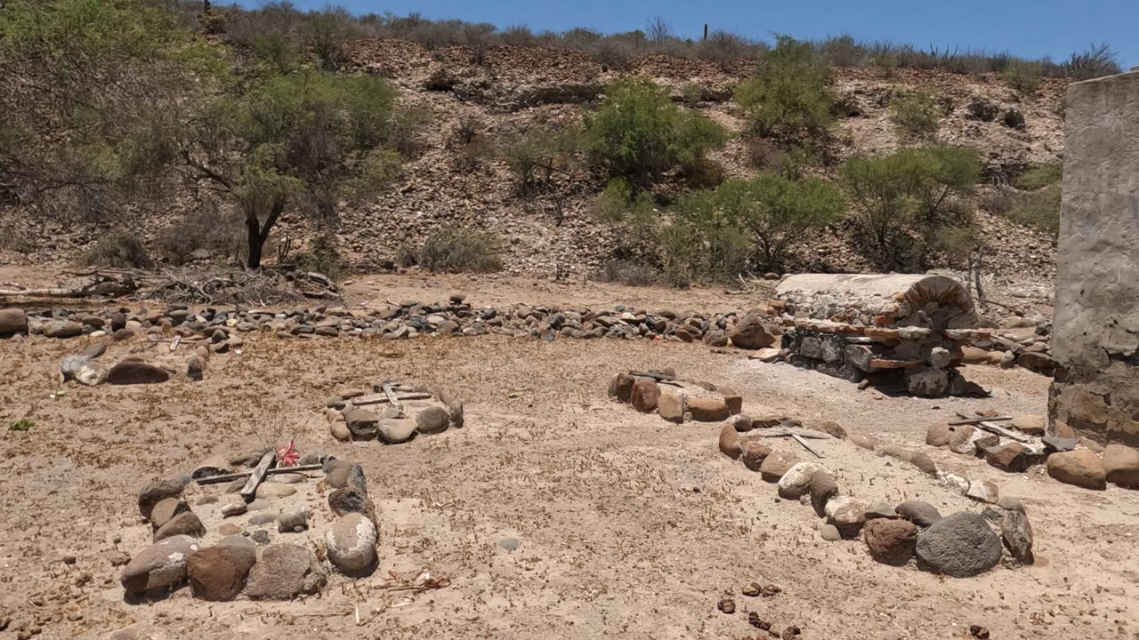 Low stone formations sit on sandy Baja California, Mexico ground, with a rock border, scattered scrub trees, hillside stones, and no visible people.