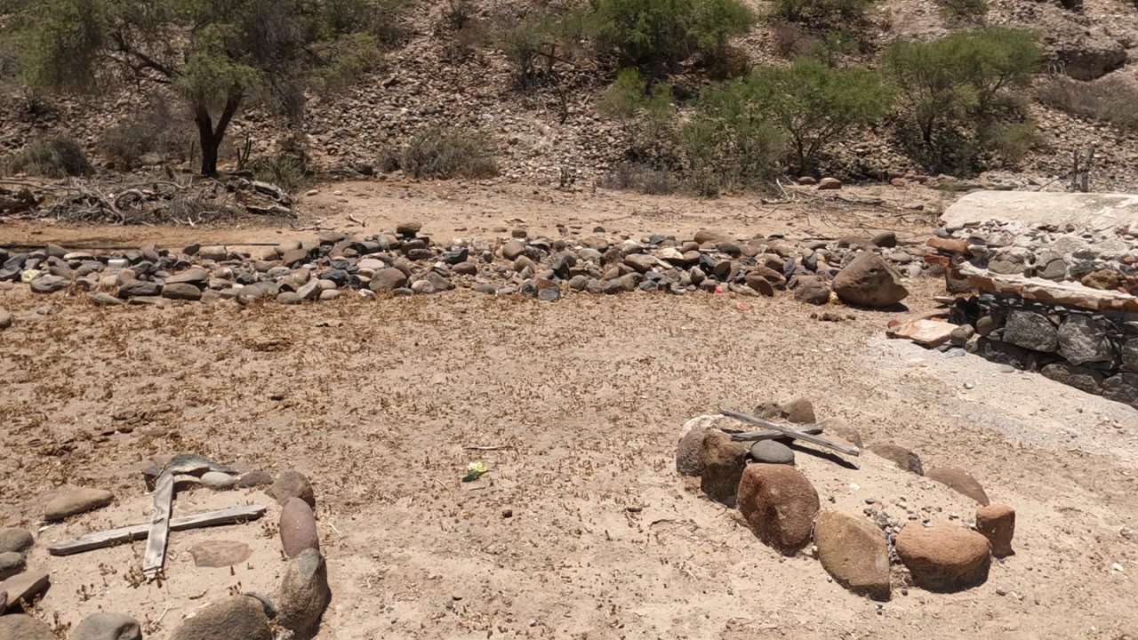 Wide view in Baja California of a sandy dry riverbed with scattered rocks, sparse trees, and a low stone and brick oven or kiln in the midground, no visible people.