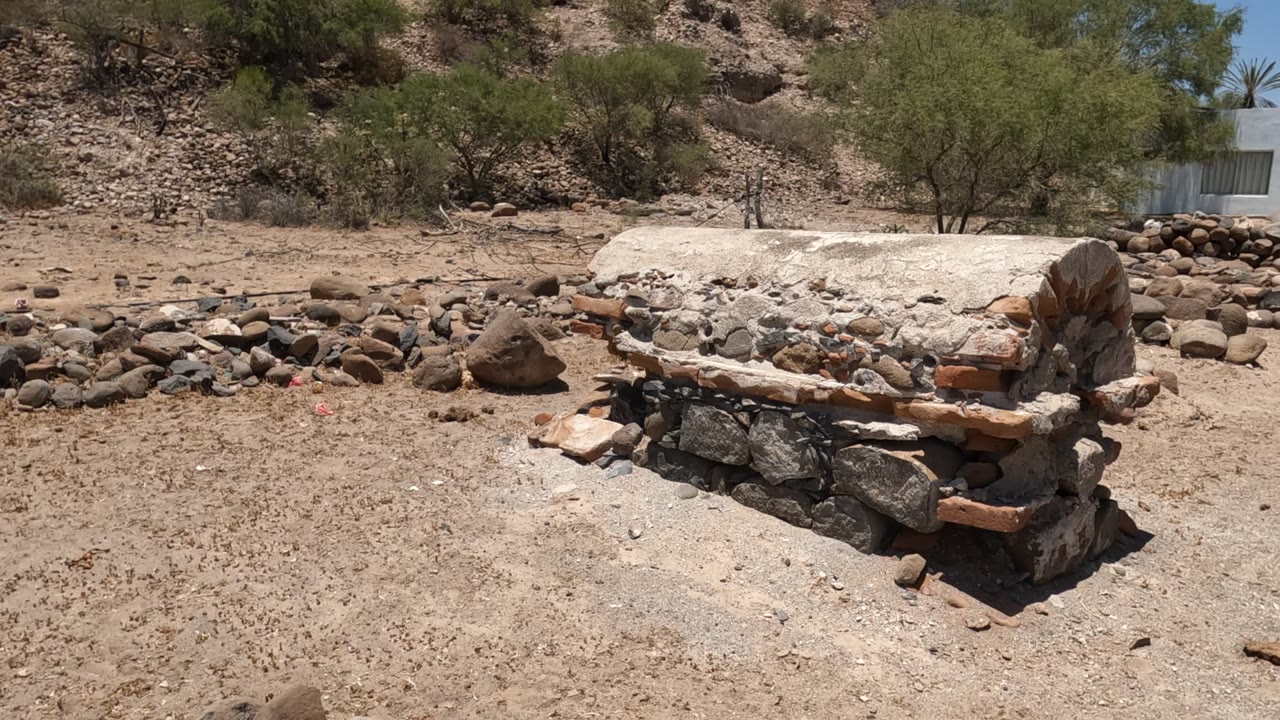 Closer side view in Baja California of the rough masonry oven or kiln with exposed brick edges, pale mortar, surrounding river stones, and sparse desert trees, no visible people.