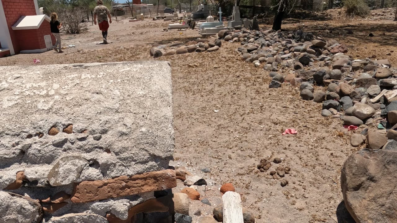 A small red building stands behind the dirt and rock construction area in Baja California, Mexico, while distant people walk near the background structures.