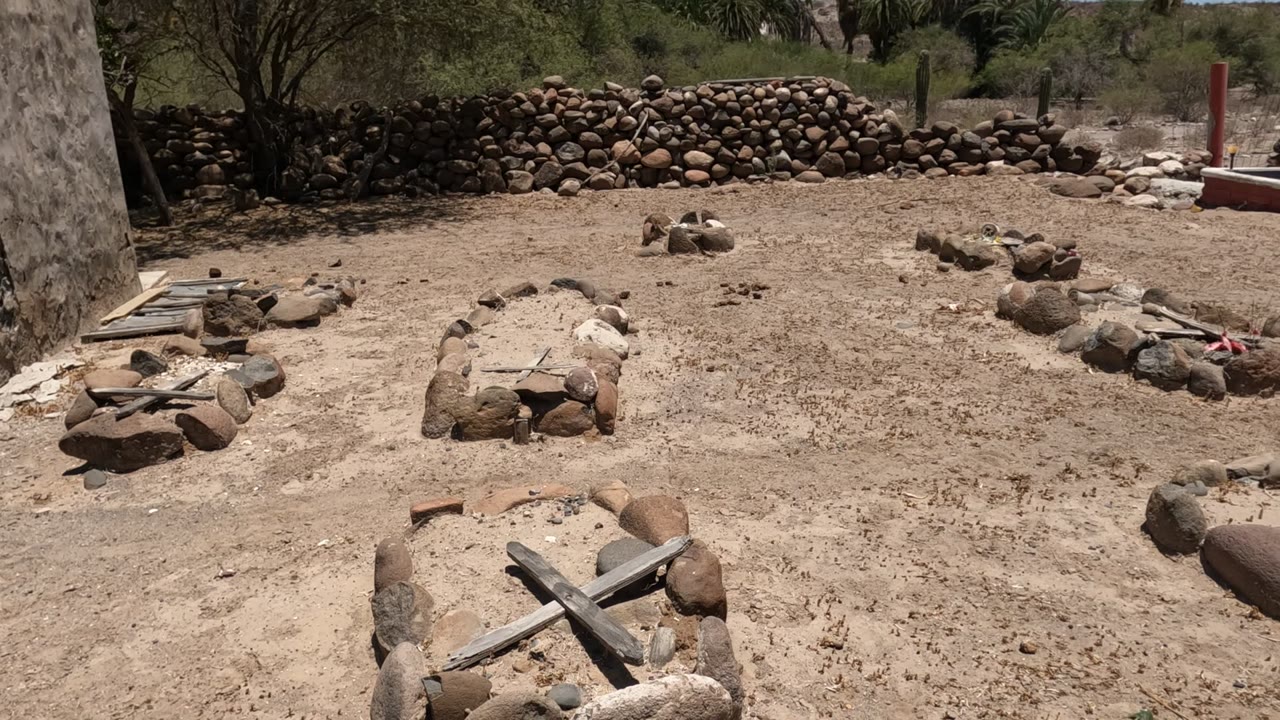 Stone grave outlines with wooden crosses sit on sandy ground in a Baja California cemetery, with a rock wall, sparse trees, palms, and no visible people.
