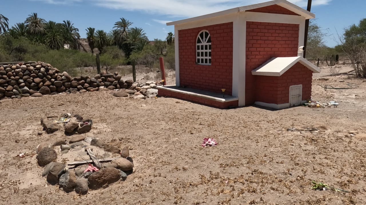 A small red brick chapel with white trim stands beside stone grave outlines and sandy ground in a Baja California cemetery, with no visible people.
