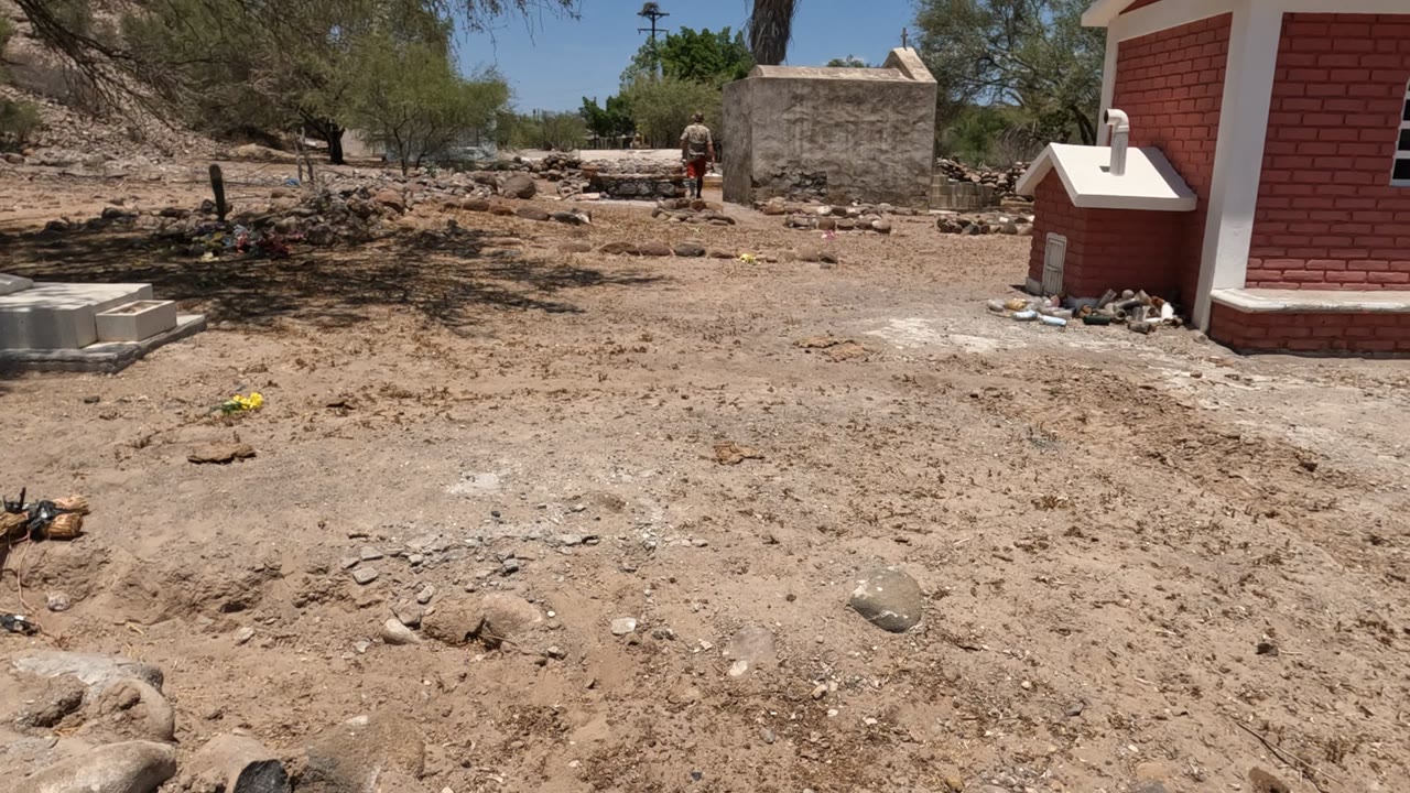 Baja California, Mexico cemetery view with sandy rocky ground, a red brick structure at right, dry trees, and a small person visible near the back wall.