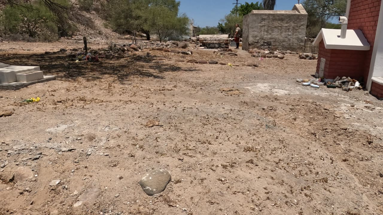 Baja California, Mexico cemetery scene with a red brick structure at the right edge, sandy foreground, dry trees, and a distant person partly visible near the rear wall.