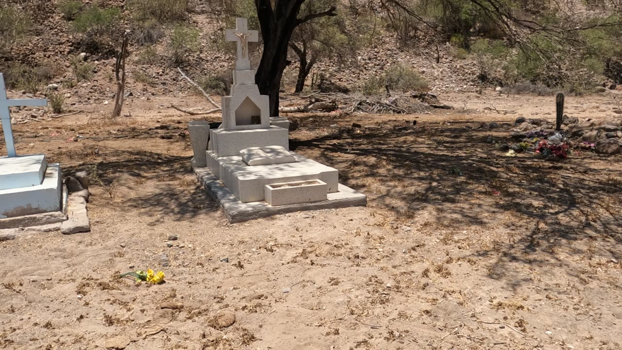 Opening frame of a Baja California desert cemetery with white cross grave markers, sandy ground, tree shade, dry hillside, and no visible people.
