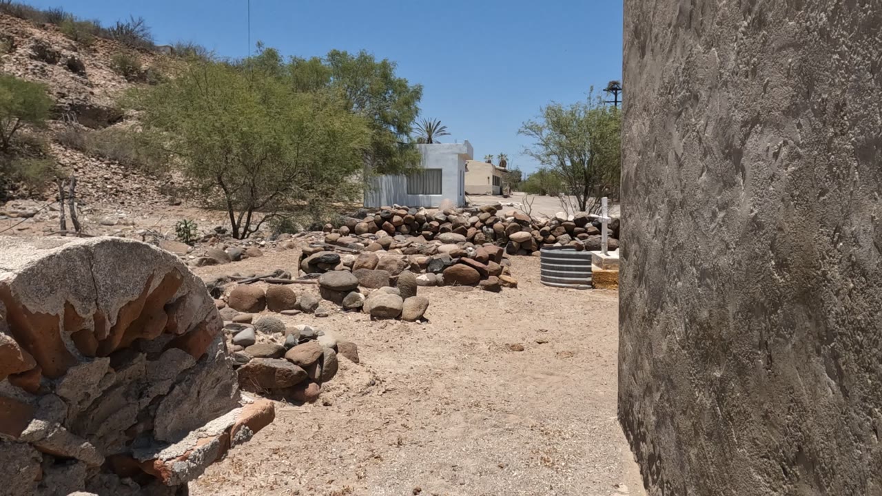 Baja California, Mexico rocky dry ground with a large stone at left, sparse trees, a pale building in the distance, and a partial person visible at the far right.