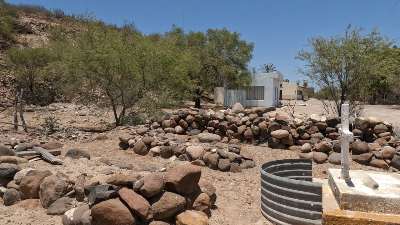 Baja California, Mexico rocky sandy yard with stacked stones, sparse trees, a white house in the background, a corrugated metal ring in the foreground, and no visible people.