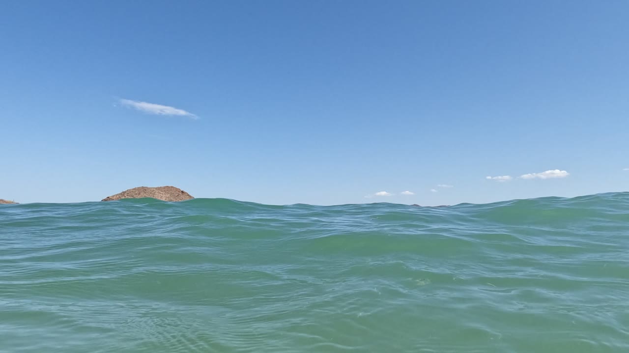 Water-level view in Baja California, Mexico, with green-blue waves in the foreground, low islands on the horizon, a clear blue sky, and no visible people.