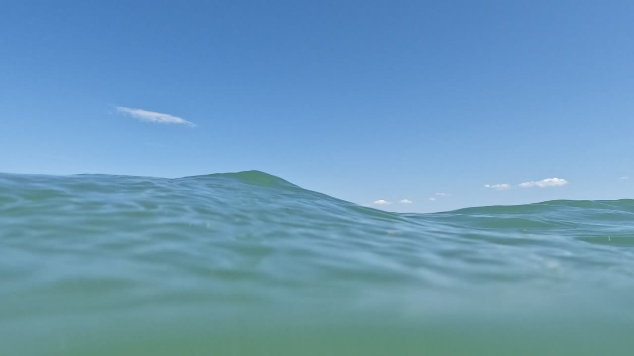 A higher foreground swell in Baja California, Mexico, partially covers the island horizon beneath a clear blue sky, with no visible people in frame.