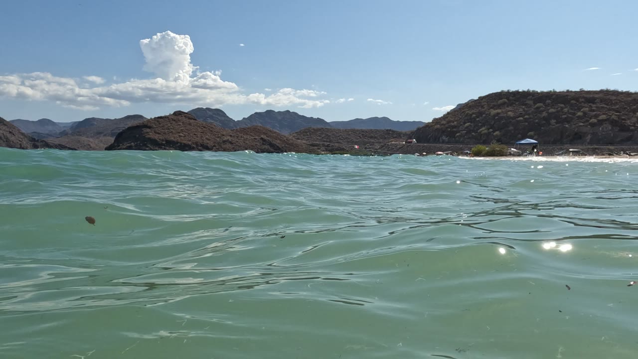 Water-level view in Baja California, Mexico with green-blue waves in the foreground, a sandy shore and umbrellas in the distance, dry hills behind them, and no visible people.