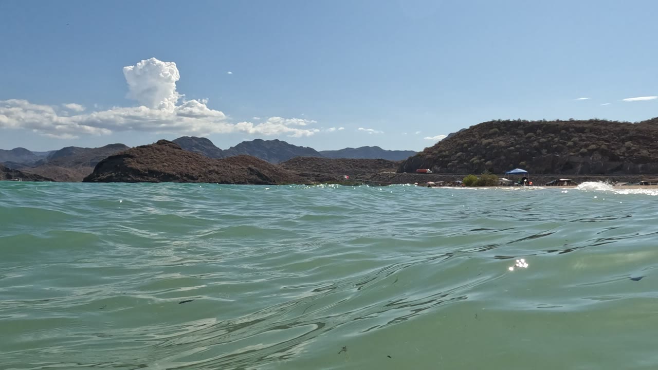 Small swells lift across the foreground in Baja California, Mexico, with a pale sandy beach, umbrellas, dark dry hills, distant mountain ridges, and no visible people.