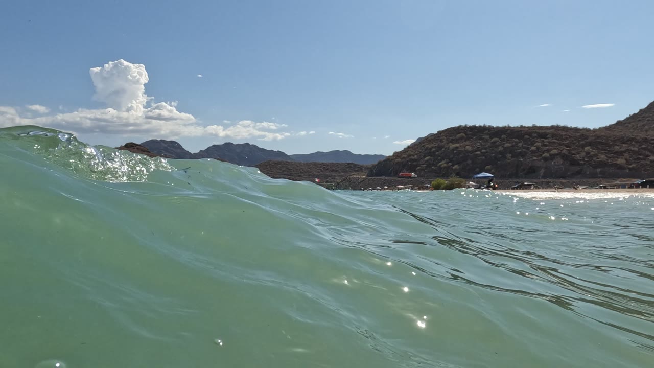 Wave crest at left with beach umbrellas and arid hills in the distance.