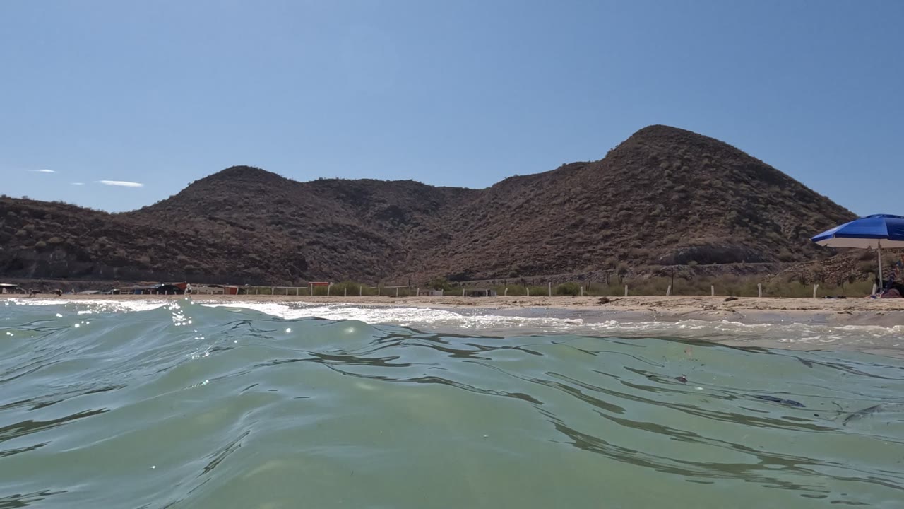 Water-level view in Baja California, Mexico with rippled green-blue ocean in the foreground, a sandy beach, dry hills under clear sky, a blue umbrella at right, and no visible people.