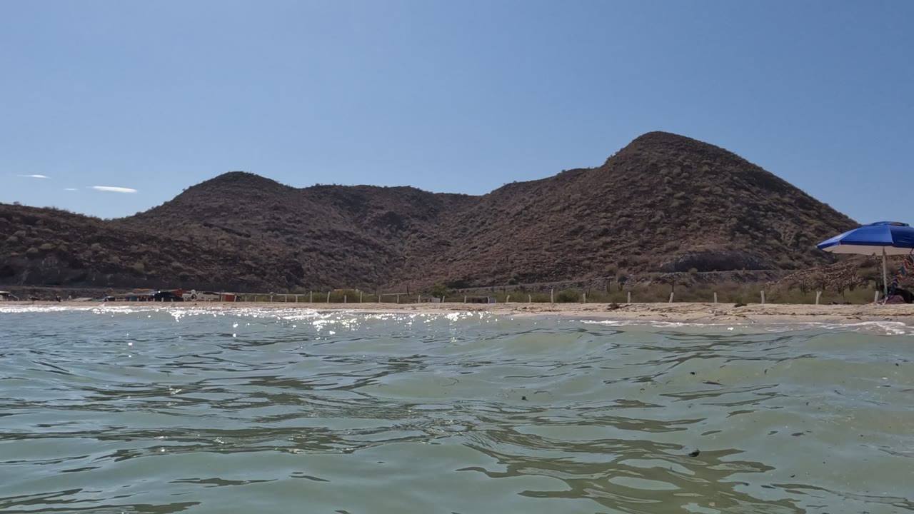 Baja California shoreline seen from low in the water, with a small wave line near the beach, dry hills across the background, clear blue sky, and no visible people.