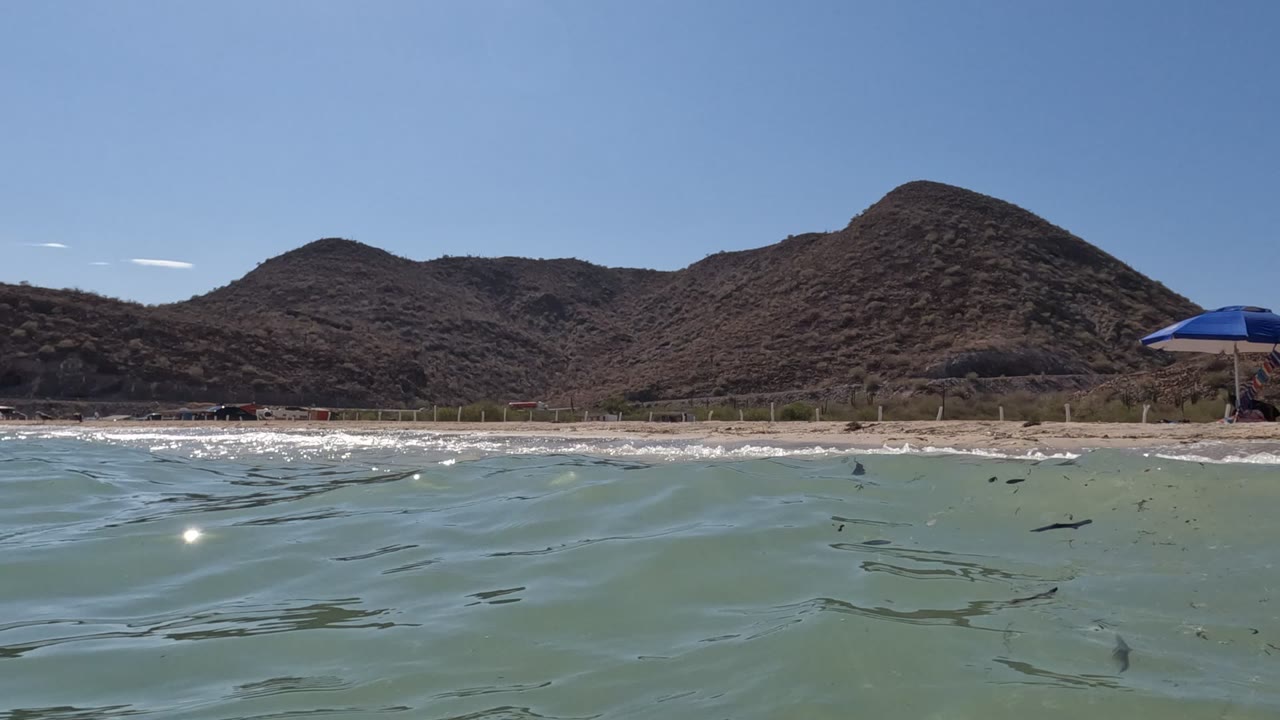 Sun glints on rippled water in Baja California, Mexico, with the sandy shore and low brown hills behind it and a blue beach umbrella near the right side, with no visible people.