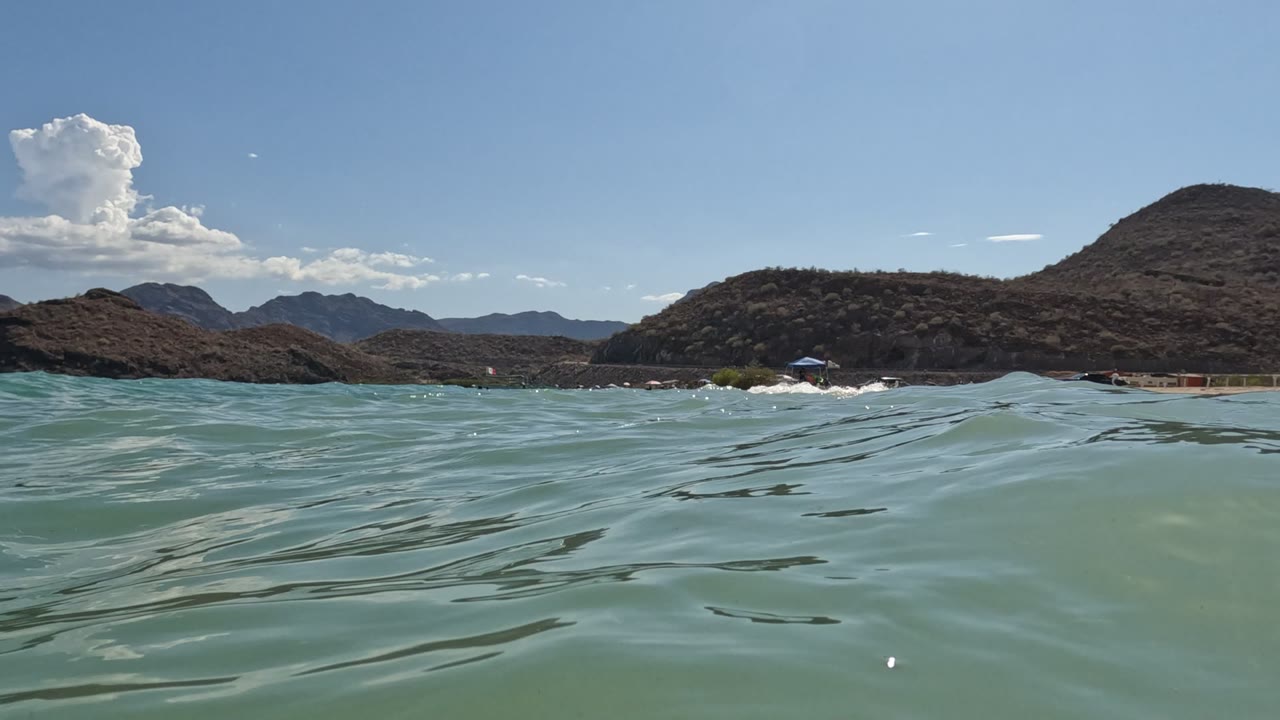 Water-level view of pale green ocean ripples facing a Baja California shoreline with rocky hills, bright sky, and no visible people.