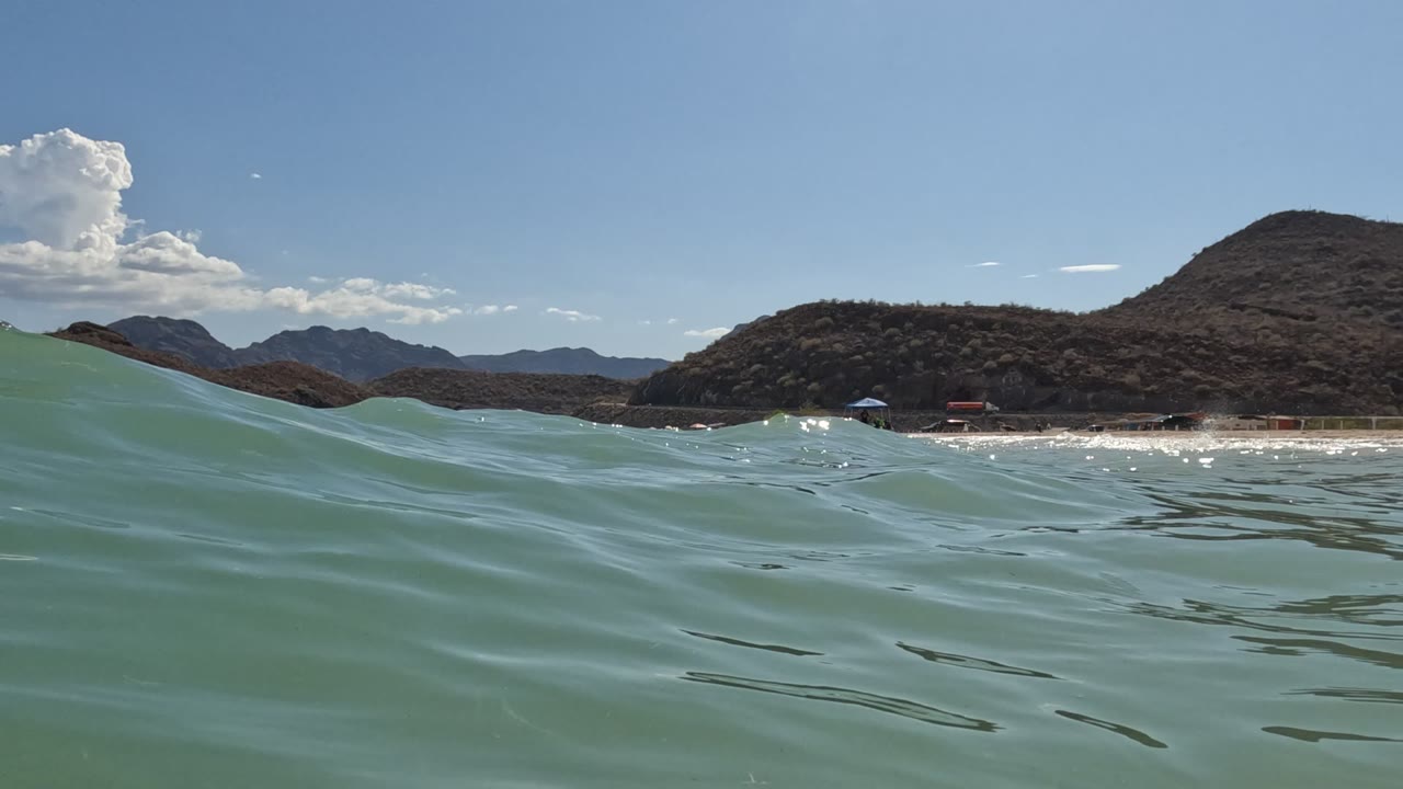 A larger foreground swell lifts across the water-level frame, partly blocking the Baja California shoreline and hills, with no visible people.