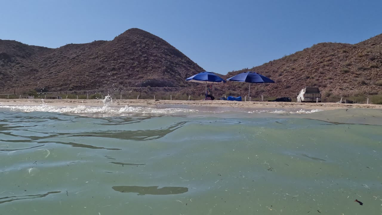Small wave splashes near the sandy shore below blue umbrellas.
