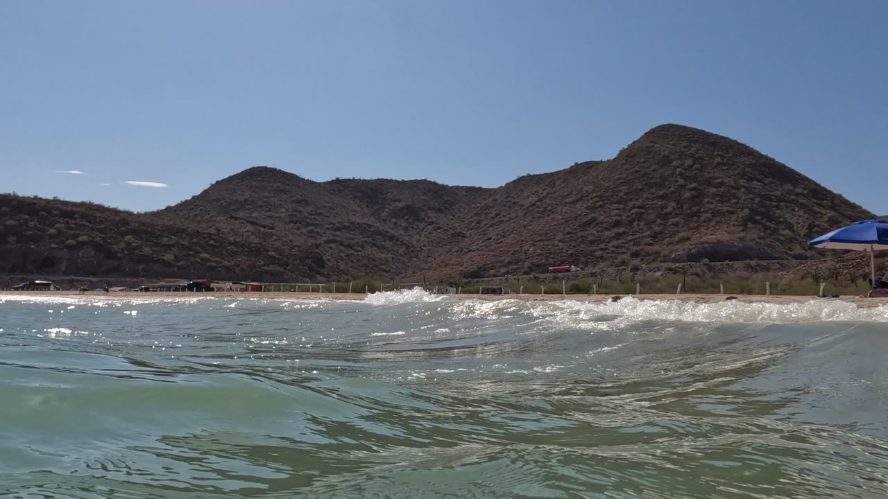 Water-level Baja California beach view with rippled ocean foreground, sandy shore and dry hills behind, a blue umbrella at the right edge, and no visible people.
