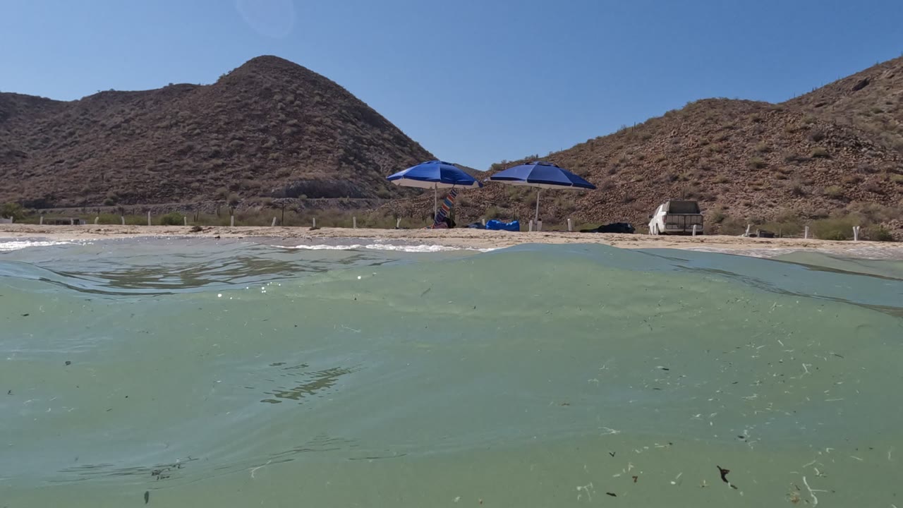 Water-level view in Baja California, Mexico, showing pale green rippled water, a sandy beach, two blue umbrellas, dry rocky hills, and no visible people.