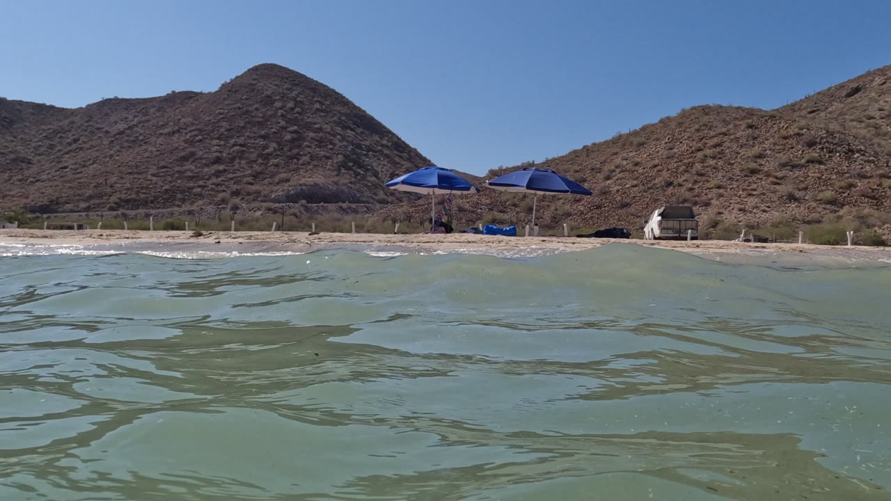 A slightly shifted ripple pattern crosses the foreground water in Baja California, Mexico, with blue umbrellas on the sand and no visible people.