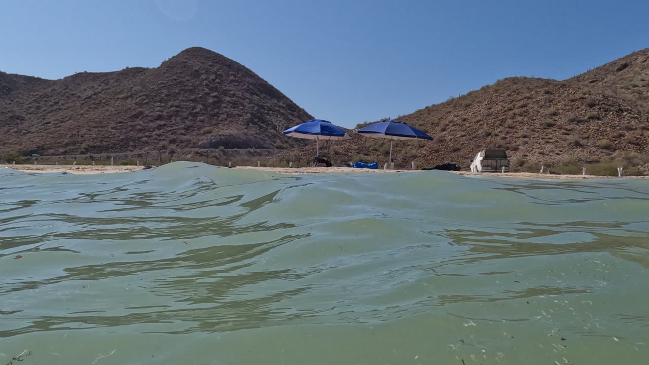 The waterline sits high in the frame as soft ripples lead toward the sandy Baja California shore, blue umbrellas, brown slopes, and no visible people.