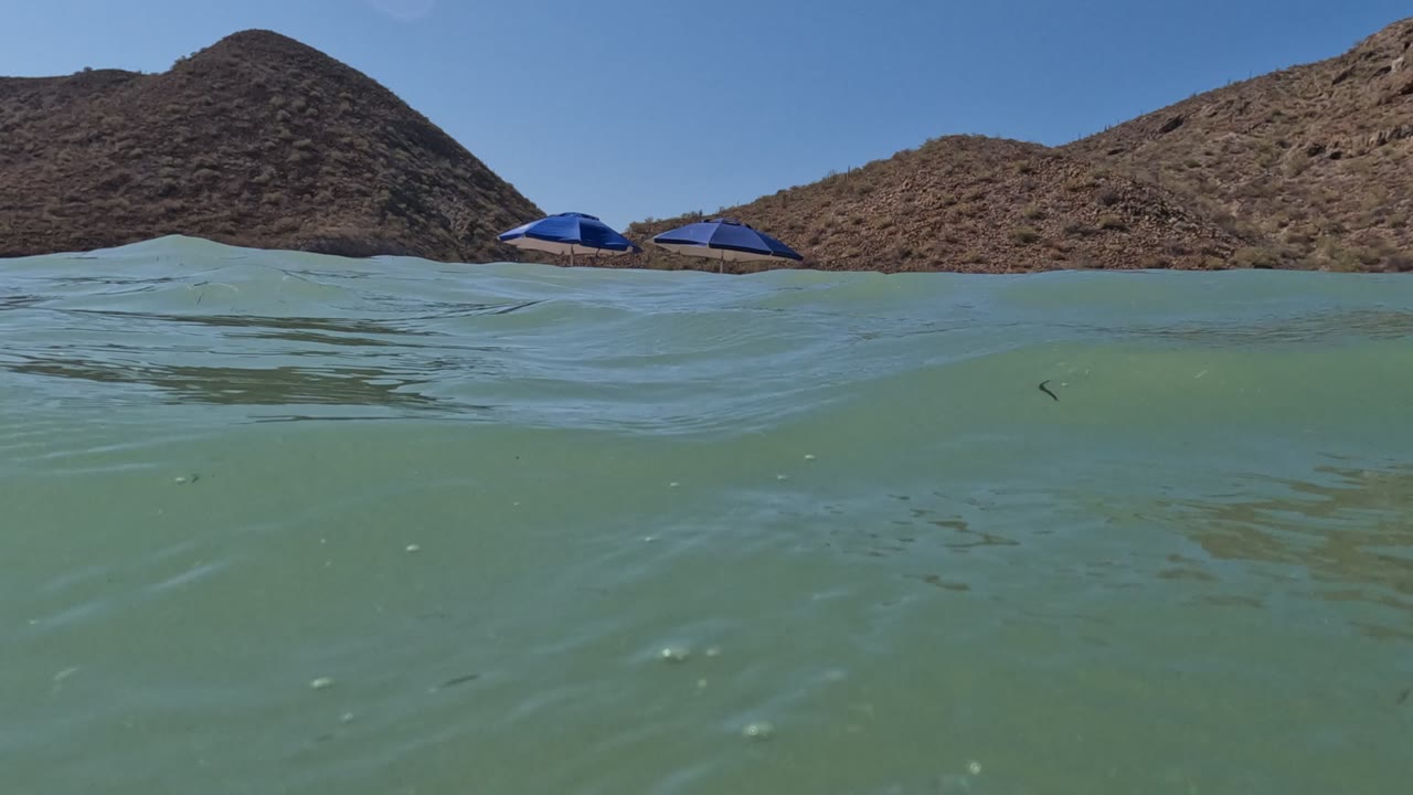 Low waterline view with blue umbrellas and hills.