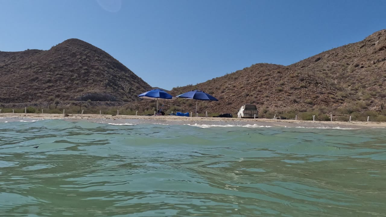 Low waterline view of beach umbrellas, sand, mountains, and blue sky.
