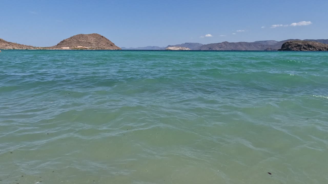 Baja California shoreline view at the start of the clip, with clear green-blue water over sand, rocky hills on the horizon, and no visible people.