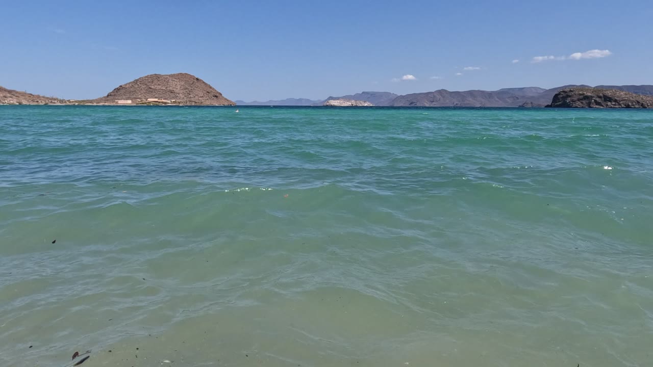 Baja California beach frame with low ripples moving across translucent water, dry hills in the background, and no visible people.