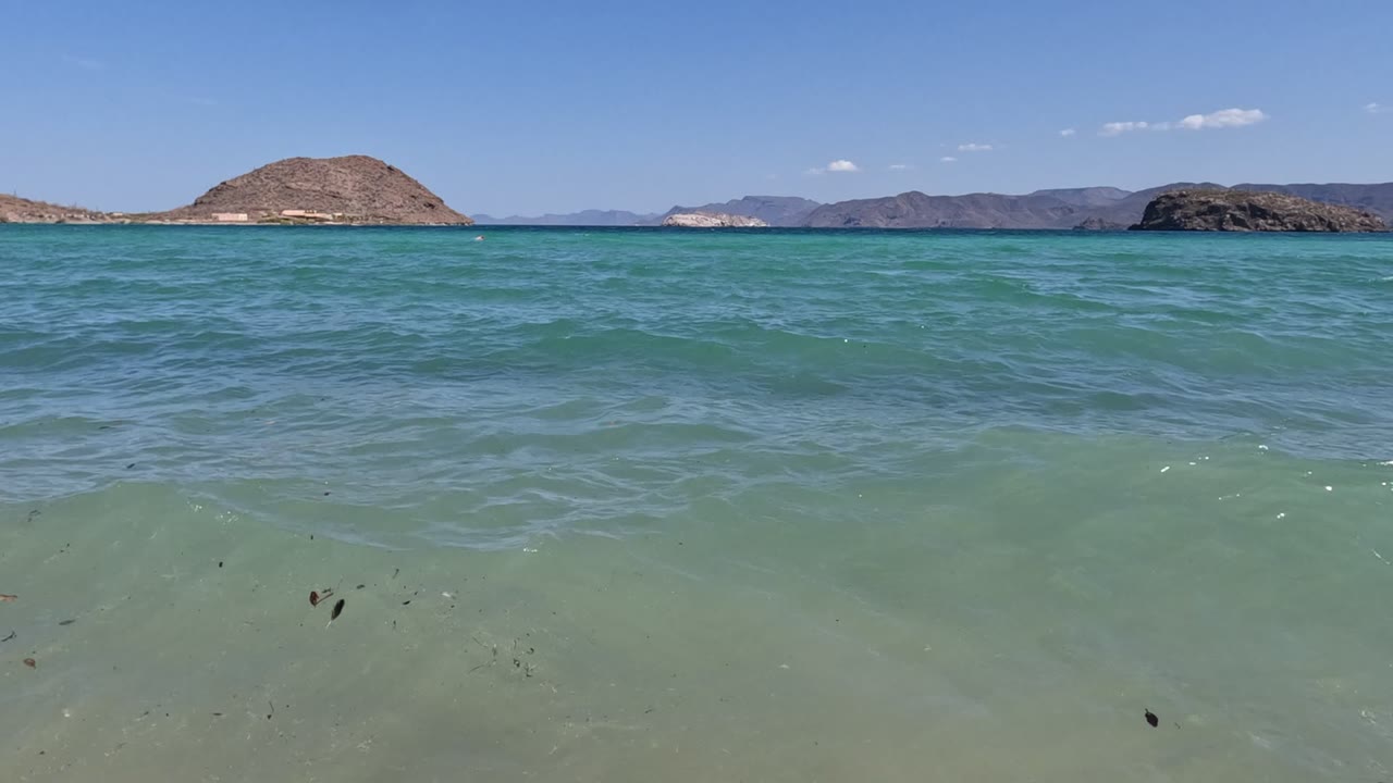Baja California coastal water fills most of the frame, with muted rocky hills and a bright blue sky beyond, and no visible people.